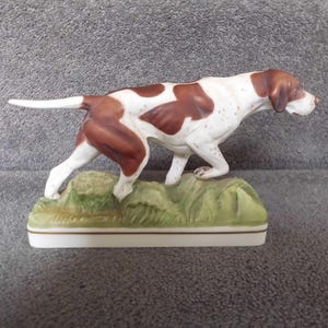 Puede incluir: Figura de porcelana de un perro en pose de caza. El perro es blanco con manchas y marcas marrones, sobre una base verde y amarilla. La figura es detallada y parece ser vintage.