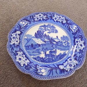May include: A round ceramic plate with a scalloped edge featuring a blue and white transferware pattern. The centre depicts a rural landscape with cottages, trees, and two deer. The border is decorated with floral motifs and a geometric rim.