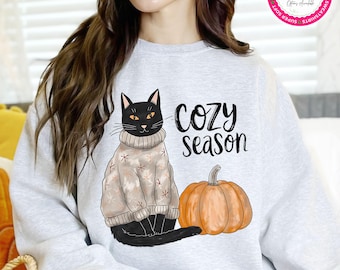 Op de afbeelding: Een lichtgrijs sweatshirt met een zwarte kat die een trui draagt en naast een oranje pompoen zit. De woorden "Cozy Season" staan boven de pompoen gedrukt. Het sweatshirt is van Bella + Canvas.