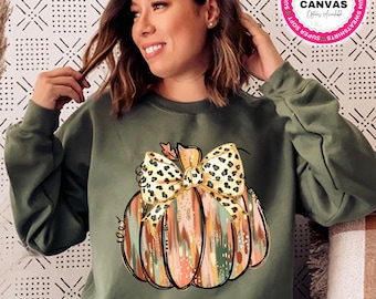 May include: A military green sweatshirt with a pumpkin design and a leopard print bow. The text "Color Pictured ~ Military Green" is at the bottom. The sweatshirt has a crew neck and long sleeves. The Bella + Canvas logo is visible.