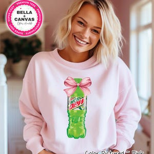 Può includere: Felpa rosa chiaro con una grafica di una bottiglia verde di Mountain Dew adornata da un fiocco rosa. Il testo "Color Pictured ~ Pink" è in basso. Il logo Bella + Canvas è visibile in alto a sinistra.