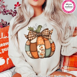 May include: A heather dust-coloured sweatshirt with a patchwork pumpkin design. The pumpkin has a brown plaid bow and is made up of various orange, brown, and floral patterned patches. The text "Color Pictured ~ Heather Dust" is at the bottom.