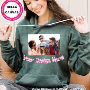 Op de afbeelding: Een bosgroene Bella + Canvas hoodie met een voorzak en een capuchon met trekkoord. De hoodie heeft een fotoprint en de tekst "Your Design Here!". De onderkant van de hoodie heeft de tekst "Color Pictured ~ Heather Forest".