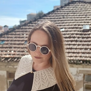 May include: A woman with long blonde hair wearing a white crocheted collar and round white sunglasses. She is standing in front of a tiled roof.
