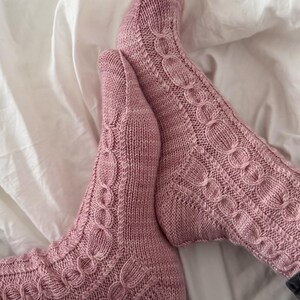Könnte beinhalten: Ein Paar gestrickte Socken in einem sanften Mauve-Ton. Die Socken haben ein Zopfmuster an den Seiten und oben, mit gerippten Bündchen und Füßen. Sie liegen auf einem weißen Laken.