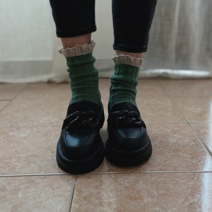 May include: Black leather loafers with a chain detail on the front. The loafers are worn with green socks that have a white lace trim.