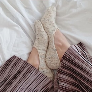 May include: A pair of beige knitted ballet socks with a subtle pattern, worn on a person's feet. The socks are on a white bed sheet with a pleated brown skirt in the foreground. The text #ballet socks is visible on the bed sheet.
