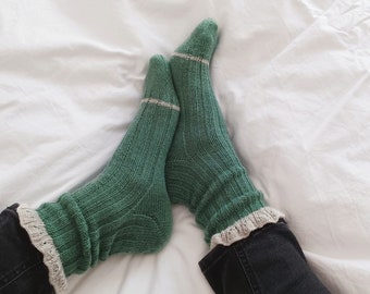 May include: A pair of green knitted socks with white lace trim. The socks are worn on a person's feet and the person is wearing black jeans.