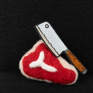 Puede incluir: Un filete de fieltro rojo con un hueso blanco y un cuchillo de carnicero bordado blanco y marrón en la parte superior.