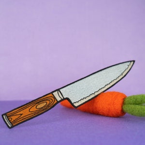 May include: A white and orange embroidered patch of a kitchen knife cutting a carrot. The knife has a brown wooden handle and a serrated blade. The carrot is orange and has a green leafy top.