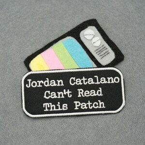 Op de afbeelding: Zwart-witte geborduurde patch met de tekst "Jordan Catalano Can't Read This Patch" en een kleurrijk retro-tv-ontwerp.