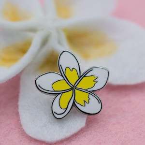 May include: A white and yellow enamel pin featuring a stylized plumeria flower.