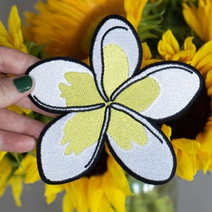 May include: A white and yellow embroidered flower patch with a black outline. The flower has five petals and a yellow center.