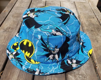 May include: A blue bucket hat with a Batman print. The hat features a repeating pattern of Batman characters and lightning bolts.