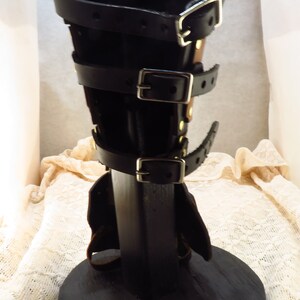 May include: Black leather leg armor with multiple buckles and straps. The armor is displayed on a black wooden stand.
