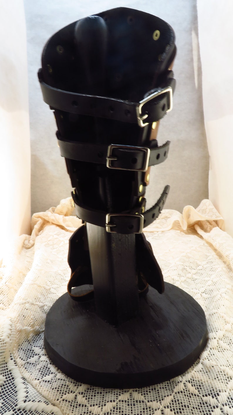 May include: Black leather leg armor with multiple buckles and straps. The armor is displayed on a black wooden stand.