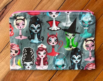 Op de afbeelding: Een grijze ritszak met een roze rits. De zak is bedekt met een kleurrijke print van verschillende cartoonfiguren met verschillende kapsels en outfits. De personages omvatten een heks, een vampier, een zombie, een buitenaardse en een verpleegster.