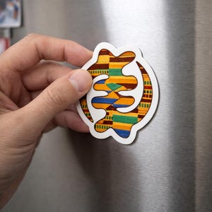 Könnte beinhalten: Ein weißer Magnet mit einem farbenfrohen Adinkra-Symbol-Design. Der Magnet zeigt einen gemusterten Stoff mit gelben, grünen, blauen und braunen Farbtönen. Der Magnet wird an einer silbernen Kühlschranktür gehalten.