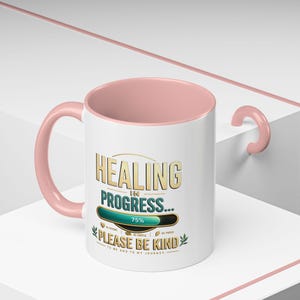 Könnte beinhalten: Weiße Keramik-Tasse mit rosa Griff und Innenraum. Die Tasse zeigt den Text "HEALING IN PROGRESS..." mit einem Fortschrittsbalken von 75 % und dem Satz "PLEASE BE KIND".