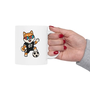 Könnte beinhalten: Ein weißer Keramikbecher mit einer Cartoon-Illustration eines Shiba Inu-Hundes, der eine bunte Sonnenbrille und ein schwarzes Trikot mit der Nummer 07 trägt und eine Pfote auf einen schwarz-weißen Fußball gestützt hat.