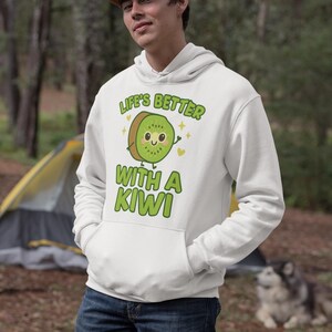 Könnte beinhalten: Weißer Kapuzenpullover mit einem Kiwi-Frucht-Motiv und dem Text "Life's Better With A Kiwi". Die Kiwi-Illustration ist grün und braun mit einem lächelnden Gesicht. Der Hoodie hat eine Kängurutasche und eine Kapuze mit Kordelzug.