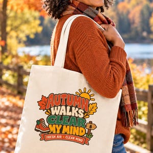 Könnte beinhalten: Eine cremefarbene Canvas-Einkaufstasche mit einem bunten Grafikdruck mit dem Text "AUTUMN WALKS CLEAR MY MIND" und "FRESH AIR • CLEAR MIND", begleitet von Illustrationen einer Sonne, Ahornblättern, einem Wanderschuh und einem hölzernen Wegweiser.