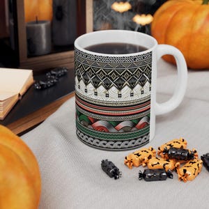 May include: A white ceramic mug with a decorative pattern in black, white, red, and green. The mug is filled with a dark liquid and sits on a light gray surface. Halloween-themed candies are scattered around the mug.