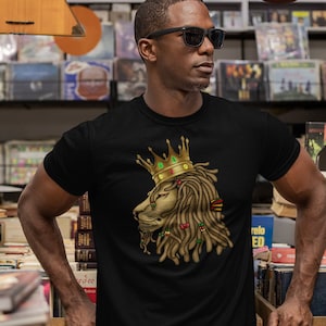 Könnte beinhalten: Ein schwarzes T-Shirt mit einem Aufdruck eines Löwen, der eine goldene Krone und Dreadlocks trägt. Die Löwenmähne ist rot, grün und gelb.