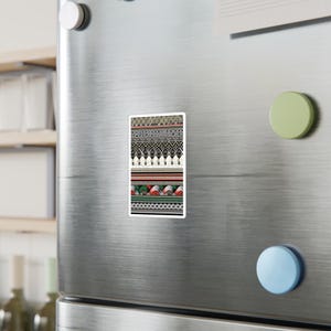 Könnte beinhalten: Ein rechteckiger Magnet mit einem farbenfrohen, geometrischen Muster. Das Design zeigt Bänder in Schwarz, Weiß, Rot und Grün mit sich wiederholenden Formen. Der Magnet ist an einer Kühlschranktür aus Edelstahl befestigt.