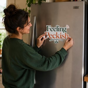 Könnte beinhalten: Ein gestanzter Vinyl-Aufkleber mit dem Text Feeling Peckish in dunkelgrüner und orangefarbener Schrift. Das Design enthält eine kleine Grafik mit Gabel und Löffel unter dem Wort Peckish sowie einen kleinen roten Sternakzent über dem Wort Feeling.