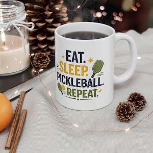 Könnte beinhalten: Ein weißer Keramikbecher mit dem in Dunkelblau, Gelb und Grün gedruckten Text EAT. SLEEP. PICKLEBALL. REPEAT. Das Design enthält kleine Abbildungen eines grünen Pickleball-Schlägers und eines gelben Balls, akzentuiert durch Sterne und Linien.