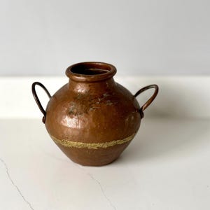 Peut inclure: Petit vase en cuivre antique avec deux anses incurvées. Le vase a un corps arrondi et un col étroit. Une bande dorée entoure le milieu du vase. Le cuivre a une patine chaude et vieillie.