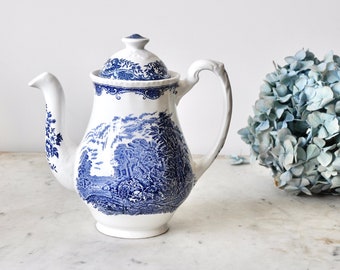 May include: A white ceramic teapot with a blue and white floral pattern. The teapot has a lid and a handle. It is sitting on a white marble surface next to a bouquet of blue hydrangeas.