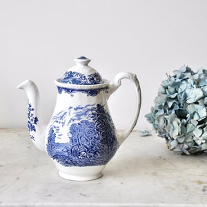 Op de afbeelding: Een witte theepot van keramiek met een blauw en wit bloemenpatroon. De theepot heeft een deksel en een handvat. Hij staat op een wit marmeren oppervlak naast een boeket blauwe hortensia's.