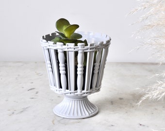 May include: White metal planter with a decorative design, featuring a small green plant inside. The planter has a pedestal base.