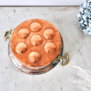 May include: A vintage copper egg poaching pan with six individual molds and two brass handles. The pan has a worn, aged patina.