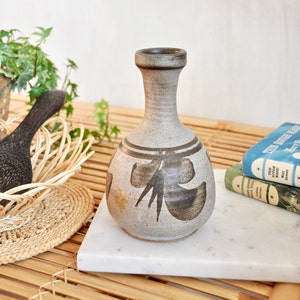 May include: A ceramic vase with a narrow neck and a bulbous body. The vase is a light beige colour with brown abstract floral designs. The vase is sitting on a white marble surface.