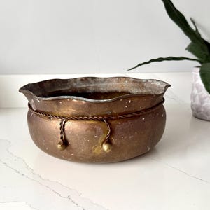 Könnte beinhalten: Eine Vintage-Schale in Kupferoptik mit gewelltem Rand und dekorativen Seildetails. Die Schale hat eine runde Form und zwei kleine messingfarbene Akzente. Die Schale wird wahrscheinlich für dekorative Zwecke oder als Pflanzgefäß verwendet.