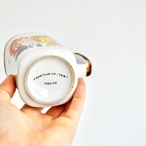 K&ouml;nnte beinhalten: Wei&szlig;e Keramiktasse mit einer Cartoon-Illustration von zwei Personen. Der Boden der Tasse tr&auml;gt den Text "CRIM Foods Ltd. - 1987 FOREIGN".