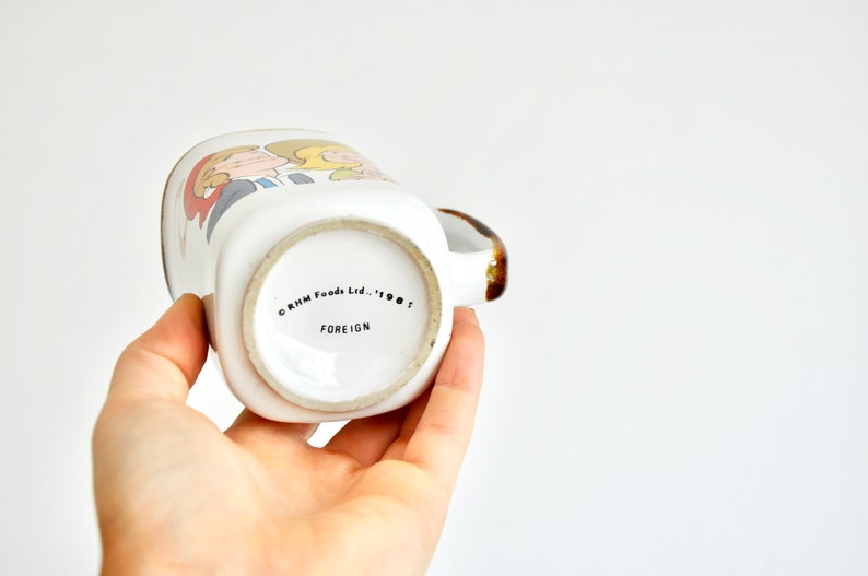 K&ouml;nnte beinhalten: Wei&szlig;e Keramiktasse mit einer Cartoon-Illustration von zwei Personen. Der Boden der Tasse tr&auml;gt den Text "CRIM Foods Ltd. - 1987 FOREIGN".