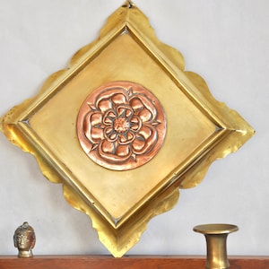 Op de afbeelding: Een vierkante messing wandplaquette met een decoratief koperen bloemmotief in het midden. De plaquette heeft een gegolfde rand en is gemonteerd op een witte muur.
