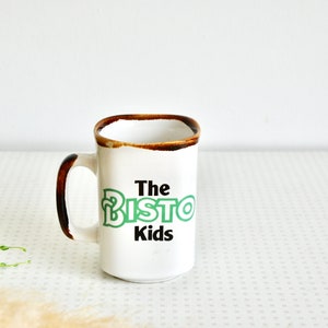 K&ouml;nnte beinhalten: Wei&szlig;e Keramiktasse mit braunem Rand und Henkel. Die Tasse hat gr&uuml;nen und schwarzen Text mit der Aufschrift "The Bisto Kids".