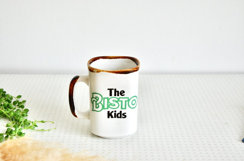 K&ouml;nnte beinhalten: Wei&szlig;e Keramiktasse mit braunem Rand und Henkel. Die Tasse hat gr&uuml;nen und schwarzen Text mit der Aufschrift "The Bisto Kids".