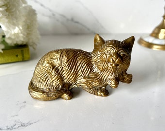 May include: A gold-colored metal figurine of a cat with a detailed, textured fur design. The cat is lying down with its head turned to the side, and its paws are tucked under its body.