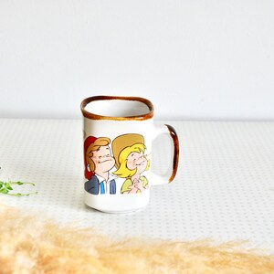 K&ouml;nnte beinhalten: Wei&szlig;e Keramiktasse mit braunem Rand und Henkel. Die Tasse zeigt eine Comic-Illustration eines Mannes und einer Frau mit H&uuml;ten.