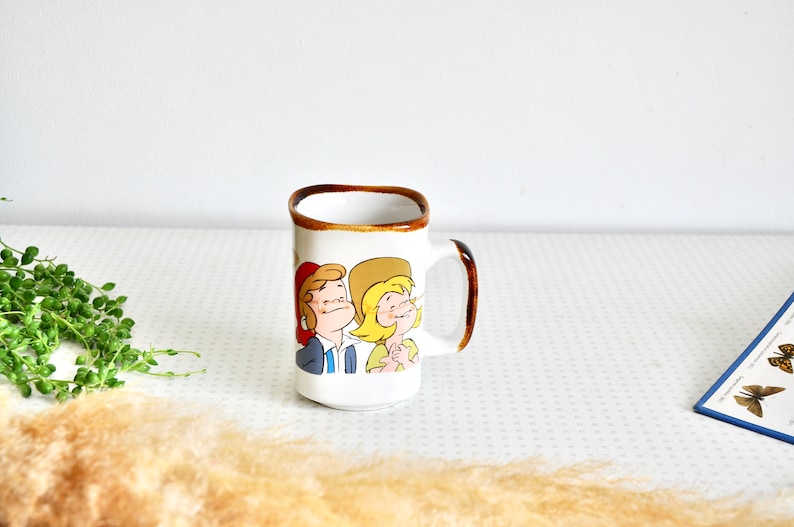 K&ouml;nnte beinhalten: Wei&szlig;e Keramiktasse mit braunem Rand und Henkel. Die Tasse zeigt eine Comic-Illustration eines Mannes und einer Frau mit H&uuml;ten.