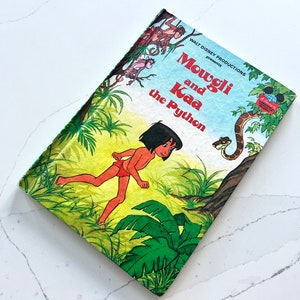 May include: A vintage children's book cover featuring Mowgli from Disney's Jungle Book. The cover depicts Mowgli running through a jungle scene with Kaa the python in the background. The title reads "Mowgli and Kaa the Python".