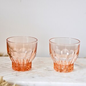 May include: Two pink glass tumblers with a textured, faceted design. The glasses are empty and sitting on a white marble surface.