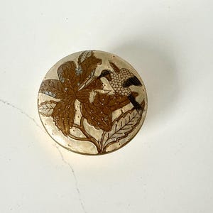 Può includere: Un portagioie rotondo e decorativo con un design floreale e di uccelli. Il coperchio presenta un'illustrazione dettagliata di un fiore marrone e un uccello con segni bianchi e neri. La scatola ha un bordo color oro e una superficie trasparente, leggermente iridescente.