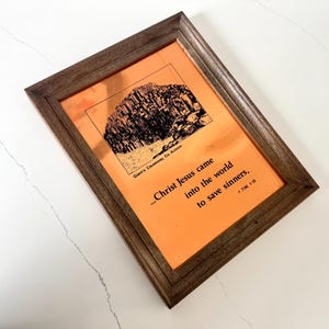 May include: Framed print with a brown wooden frame. The print has an orange background with a black and white illustration of a rock formation. Text on the print reads "...Christ Jesus came into the world to save sinners."